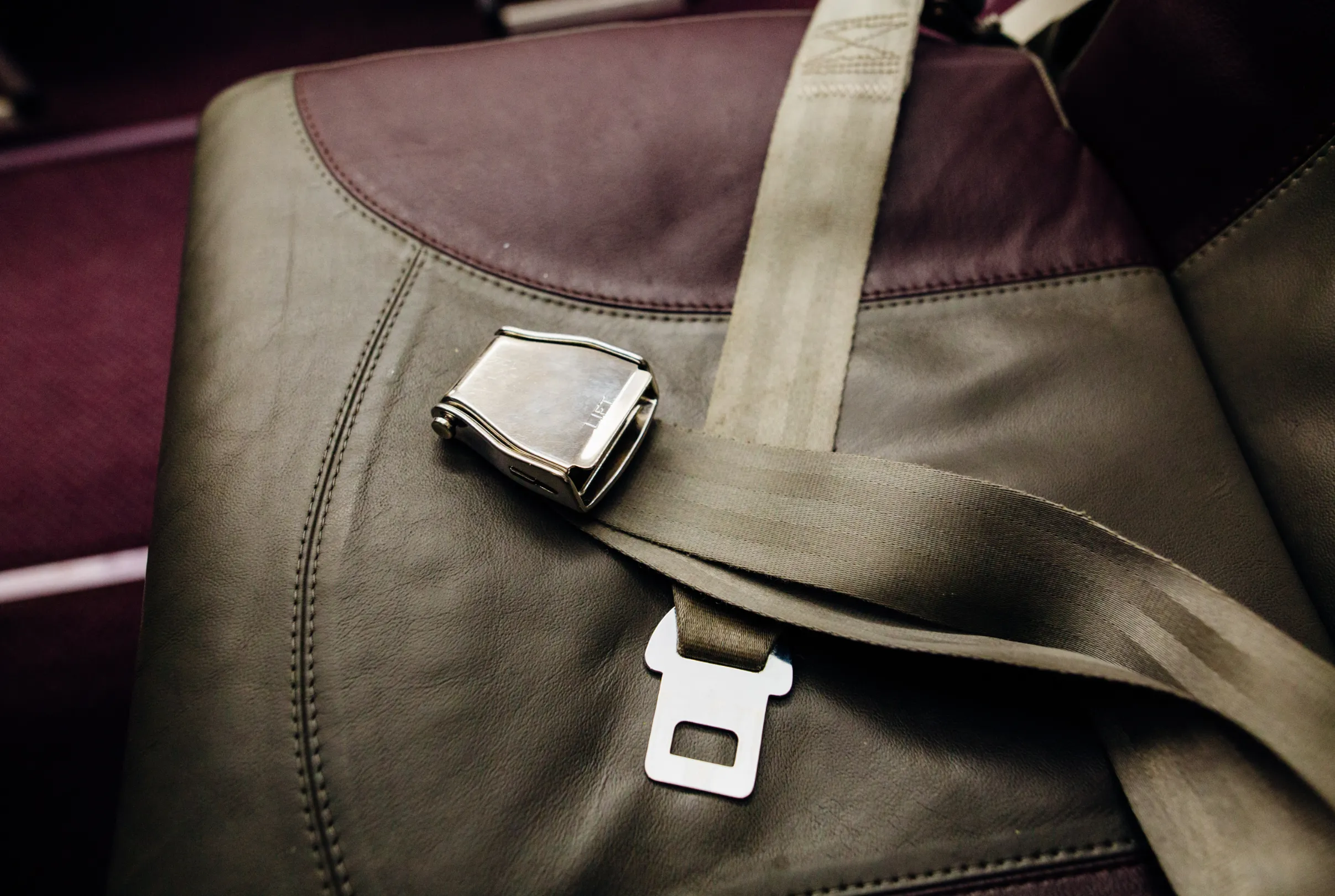 A khaki lap belt with a polished metal lift-latch buckle lying across a leather transport seat.