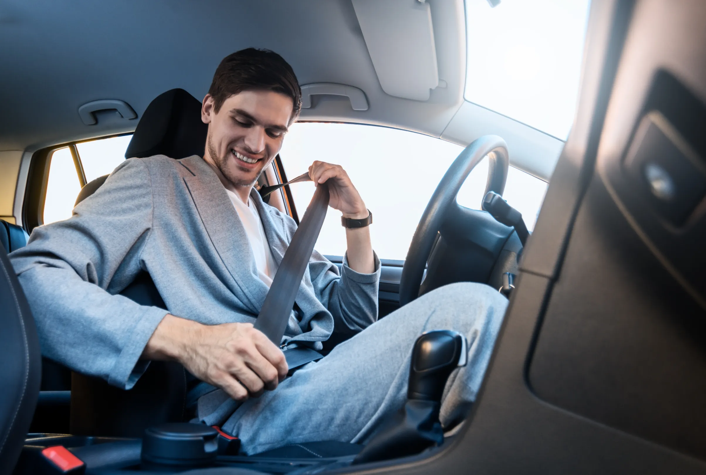 A driver seated in a car, smiling and drawing the seat belt webbing across the body before fastening it.