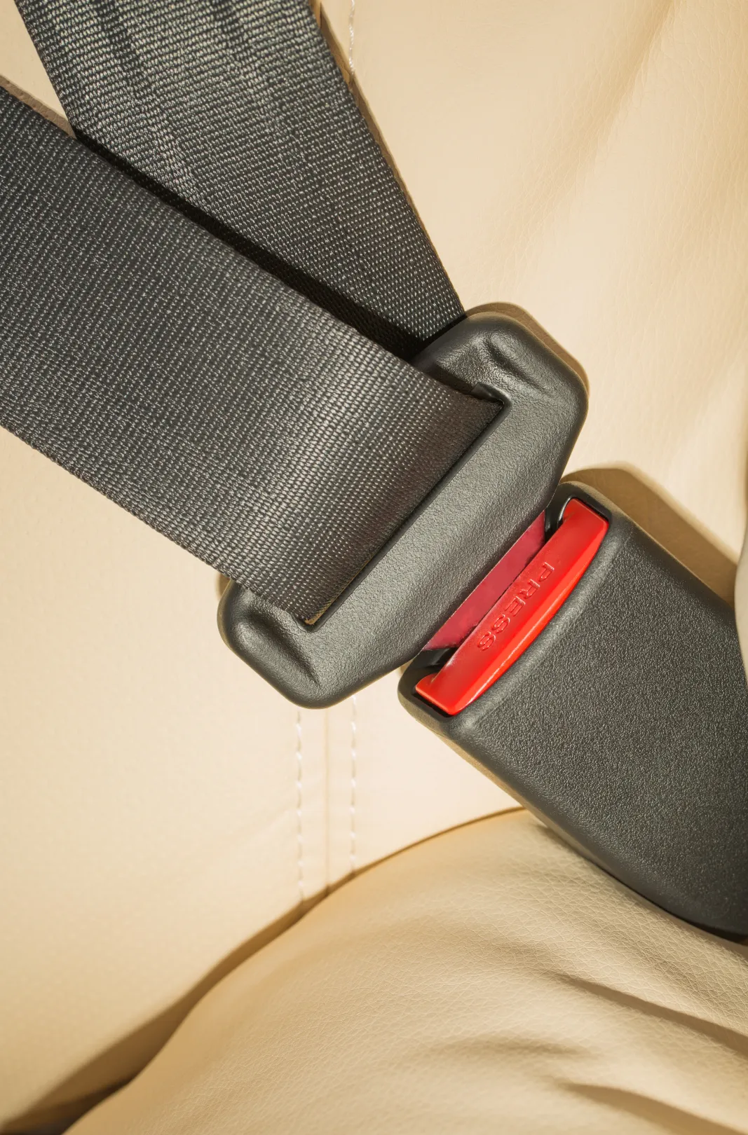 A dark seat belt webbing strap and its tongue plate latched into a buckle, photographed against a cream leather vehicle seat.