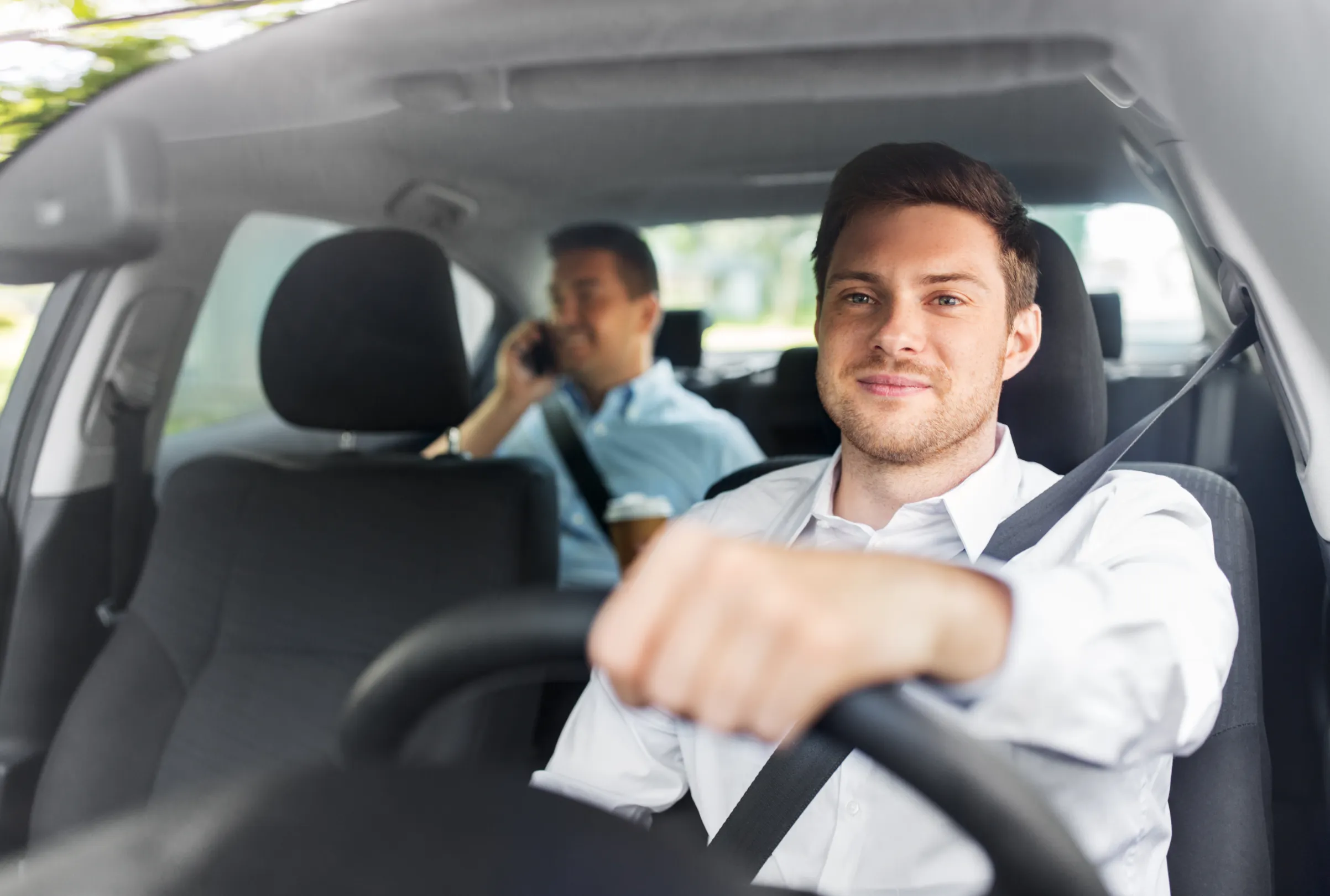 A driver in a white shirt wearing a fastened three-point seat belt with both hands on the steering wheel, a passenger seated behind.