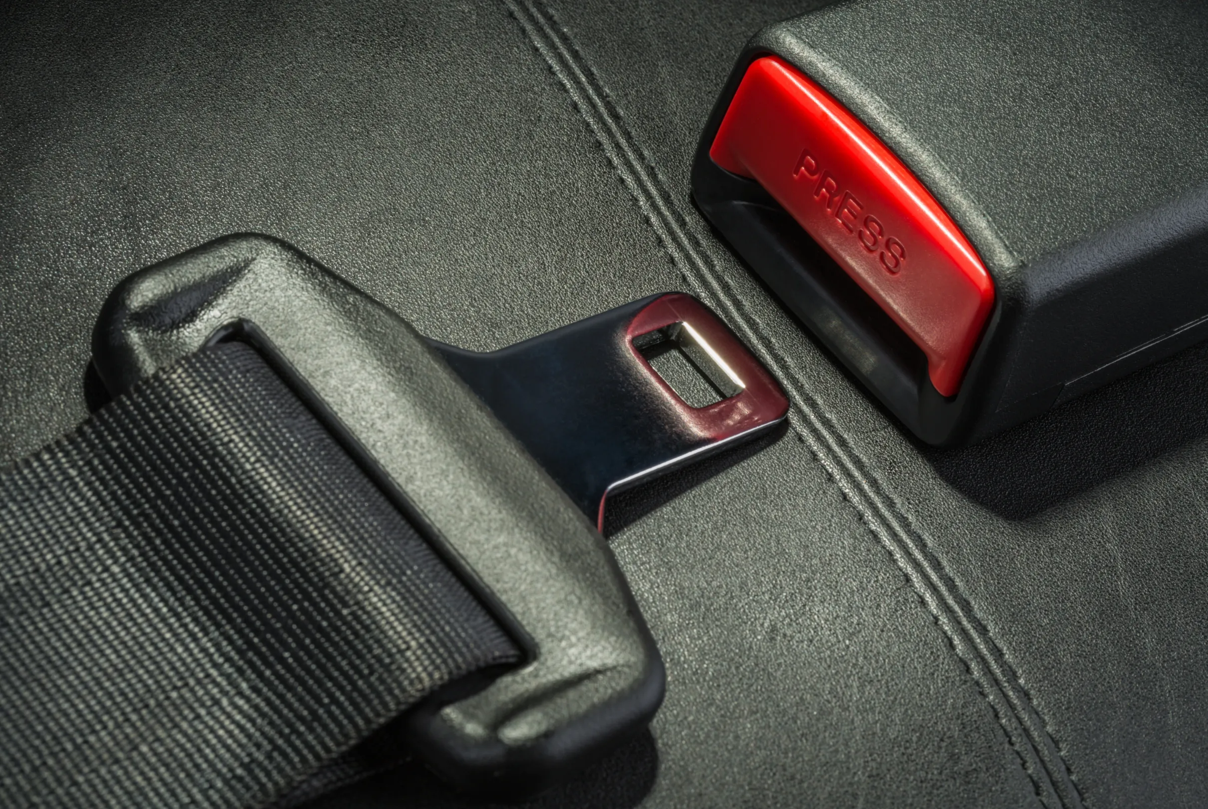 Studio close-up of a seat belt tongue plate lying beside an unfastened buckle receptacle with a red PRESS release button on black upholstery.