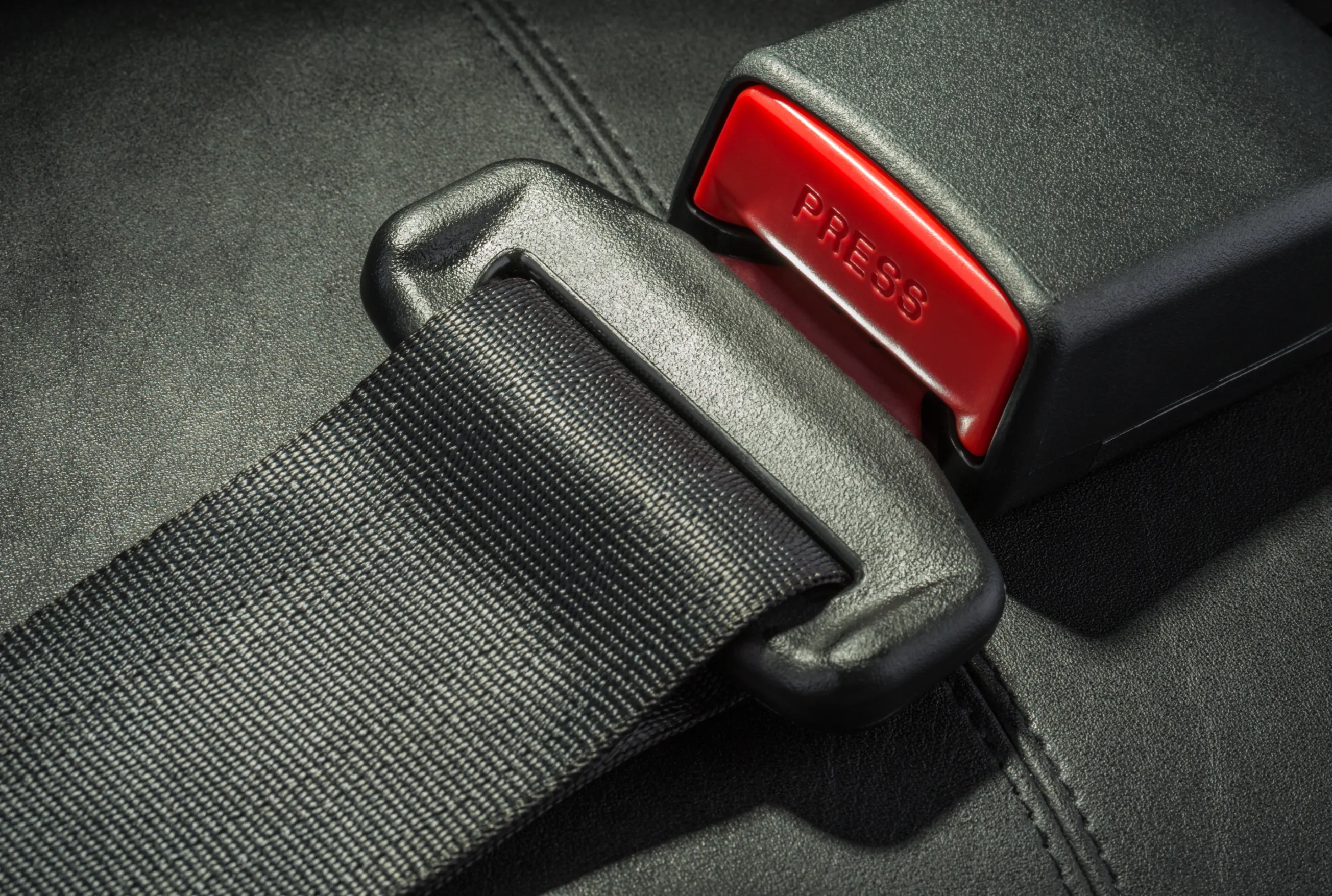 Macro photograph of a seat belt tongue plate fully latched into a black buckle receptacle with a red PRESS release button, on dark leather upholstery.