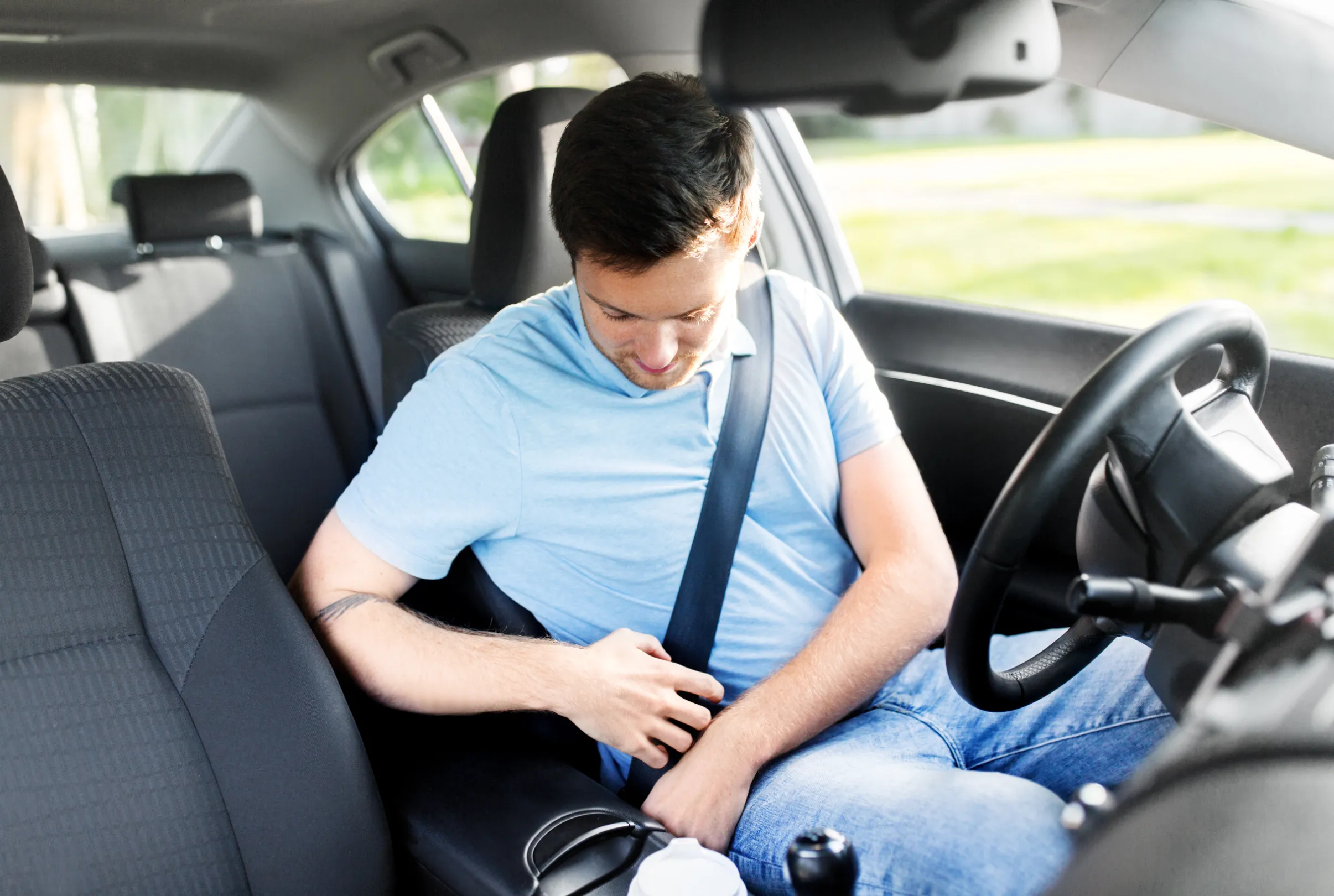 A driver seated behind the wheel securing a fastened three-point seat belt, photographed from the rear seat of the car.