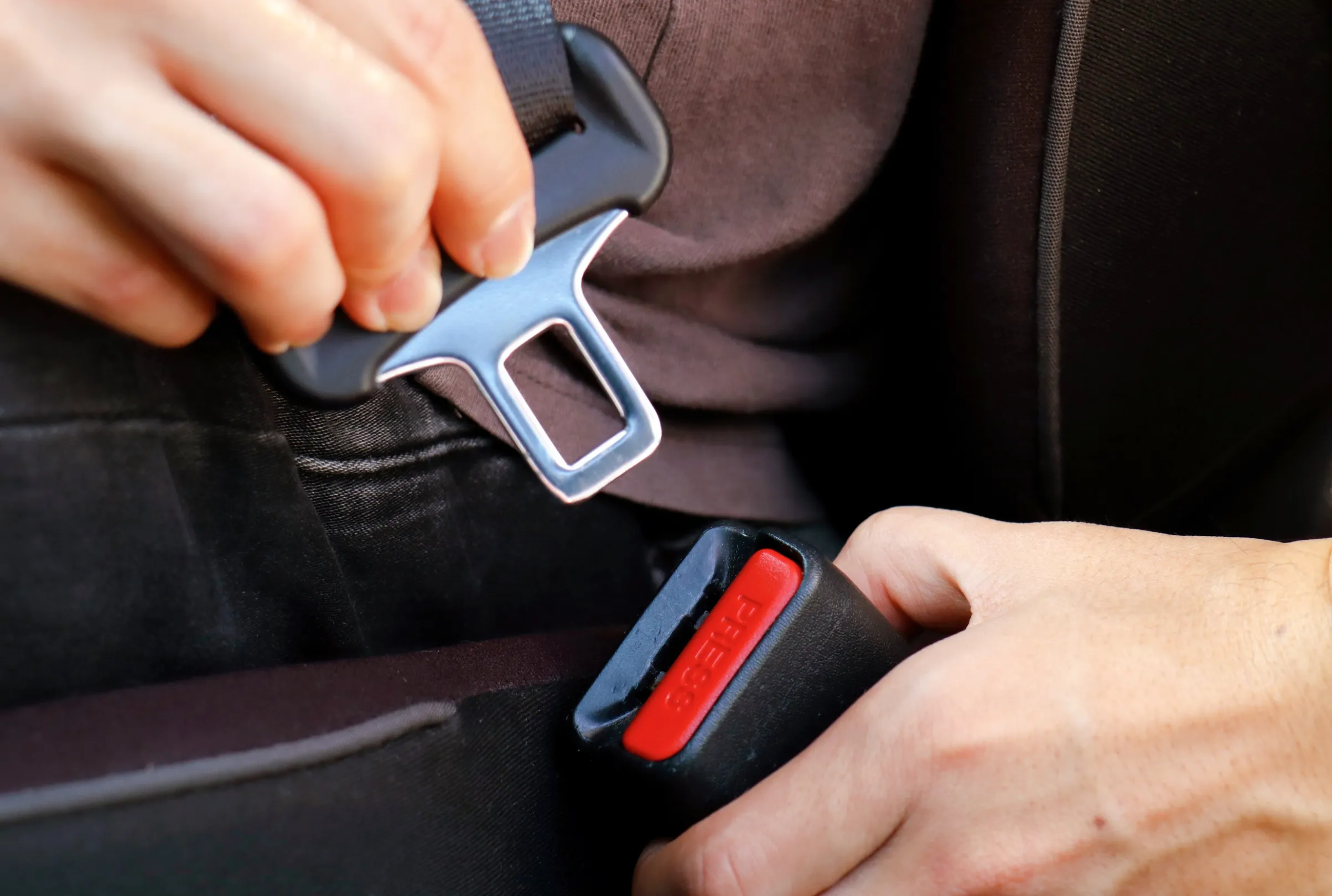 Two hands guiding a metal seat belt tongue plate towards a black buckle with a red release button, seen close up inside a vehicle.